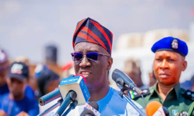 Man wearing a red-and-navy traditional cap and sunglasses speaks into multiple microphones at a press conference, with a uniformed officer in a blue beret behind him.