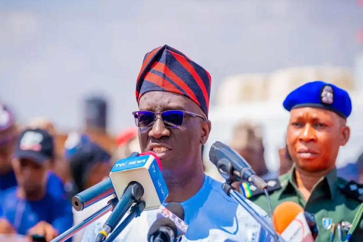 Man wearing a red-and-navy traditional cap and sunglasses speaks into multiple microphones at a press conference, with a uniformed officer in a blue beret behind him.