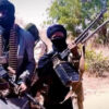 Group of masked men in military-style clothing holding rifles in a dry outdoor area on a sunny day.