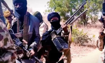 Group of masked men in military-style clothing holding rifles in a dry outdoor area on a sunny day.