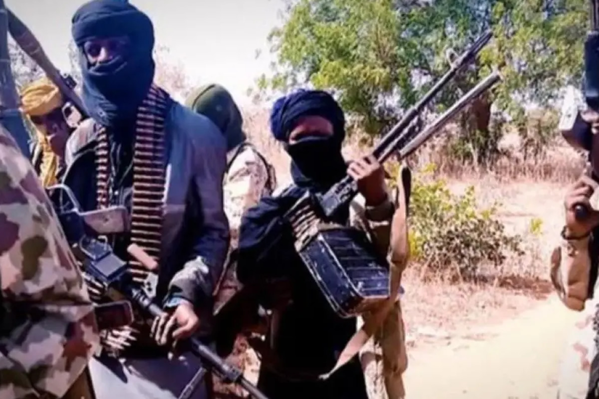 Group of masked men in military-style clothing holding rifles in a dry outdoor area on a sunny day.