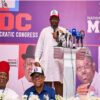 Man in white traditional attire speaks at a podium with multiple microphones, NDC banner in purple backdrop behind him.