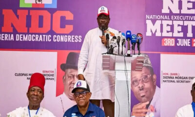 Man in white traditional attire speaks at a podium with multiple microphones, NDC banner in purple backdrop behind him.