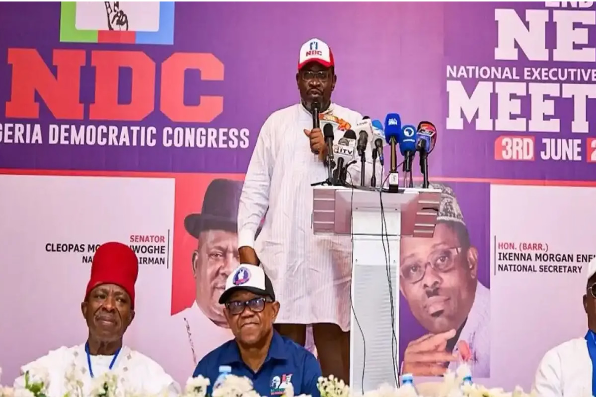Man in white traditional attire speaks at a podium with multiple microphones, NDC banner in purple backdrop behind him.