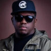 Portrait of a man facing the camera, wearing a dark cap with a white logo, black sunglasses, and a green military-style jacket against a brown background.