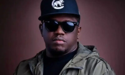 Portrait of a man facing the camera, wearing a dark cap with a white logo, black sunglasses, and a green military-style jacket against a brown background.