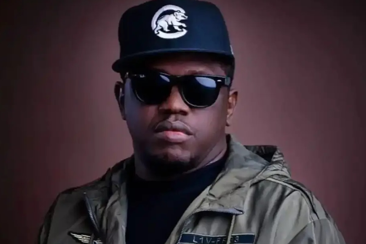 Portrait of a man facing the camera, wearing a dark cap with a white logo, black sunglasses, and a green military-style jacket against a brown background.