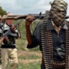 Masked group of gunmen walking through a rural field, rifles raised; front man has a scarf over his face and an ammo belt.