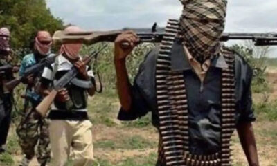 Masked group of gunmen walking through a rural field, rifles raised; front man has a scarf over his face and an ammo belt.