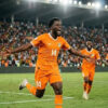 Soccer player in orange jersey #14 celebrates a goal with arms outstretched as teammates run toward him; crowded stadium in the background.