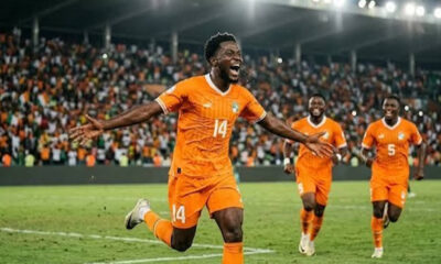 Soccer player in orange jersey #14 celebrates a goal with arms outstretched as teammates run toward him; crowded stadium in the background.