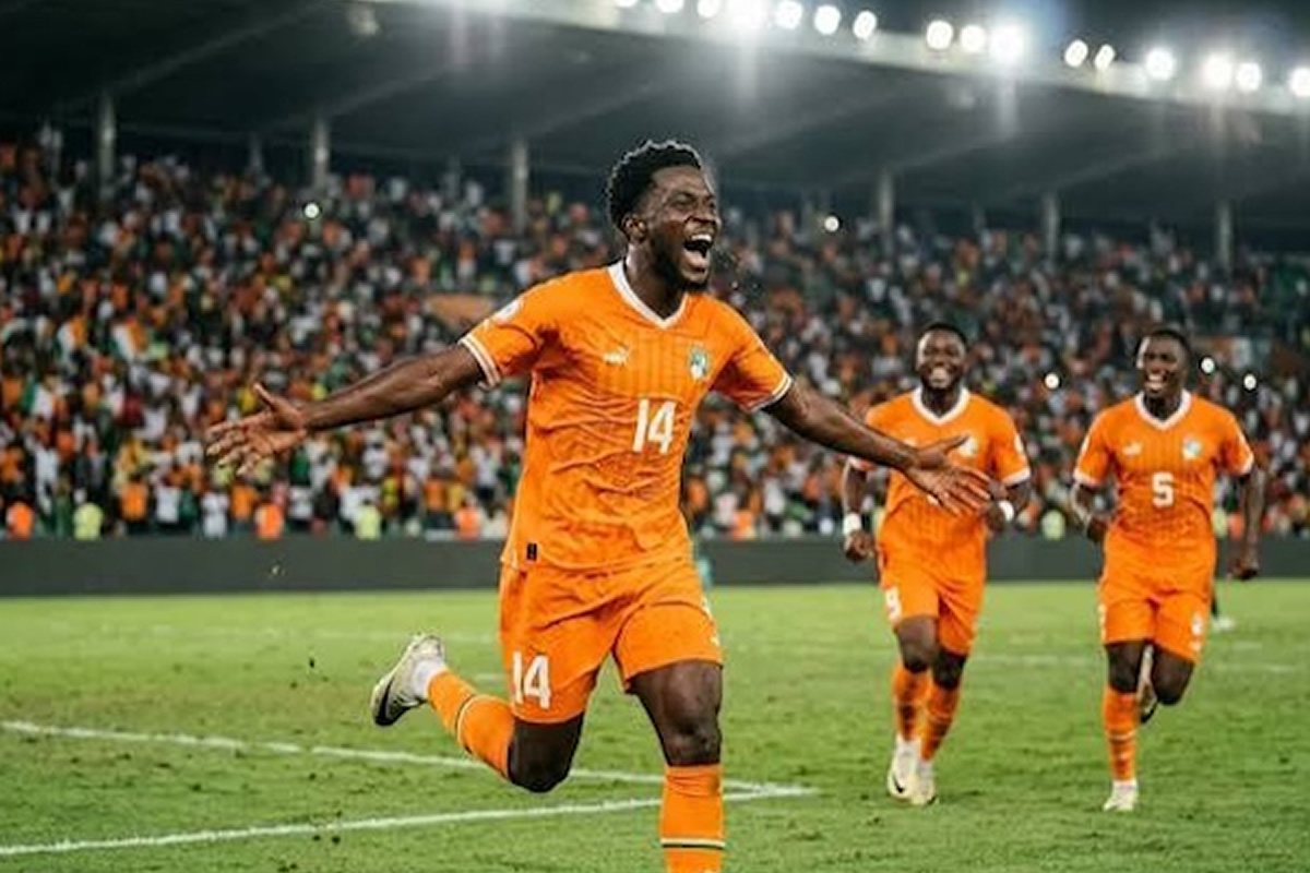 Soccer player in orange jersey #14 celebrates a goal with arms outstretched as teammates run toward him; crowded stadium in the background.
