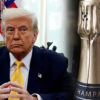 Former President Donald Trump in a navy suit and yellow tie, hands folded, with the U.S. flag in the background.