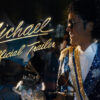 Man dressed as Michael Jackson in a blue sequined jacket smiles in the foreground with neon sign reading 'Michael Official Trailer' in the background.