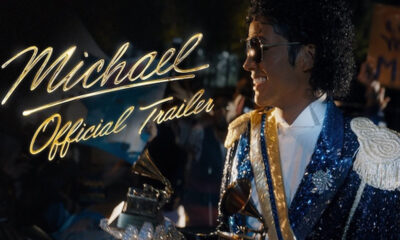 Man dressed as Michael Jackson in a blue sequined jacket smiles in the foreground with neon sign reading 'Michael Official Trailer' in the background.