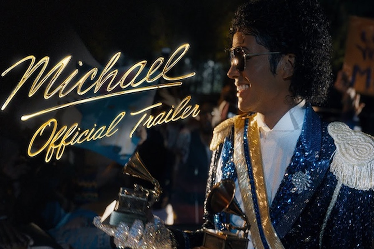 Man dressed as Michael Jackson in a blue sequined jacket smiles in the foreground with neon sign reading 'Michael Official Trailer' in the background.