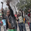 Group of men protesting in a street; a man in a black jacket raises a machete and his hand toward the camera while others hold sticks, with smoke rising in the background.