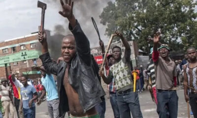Group of men protesting in a street; a man in a black jacket raises a machete and his hand toward the camera while others hold sticks, with smoke rising in the background.