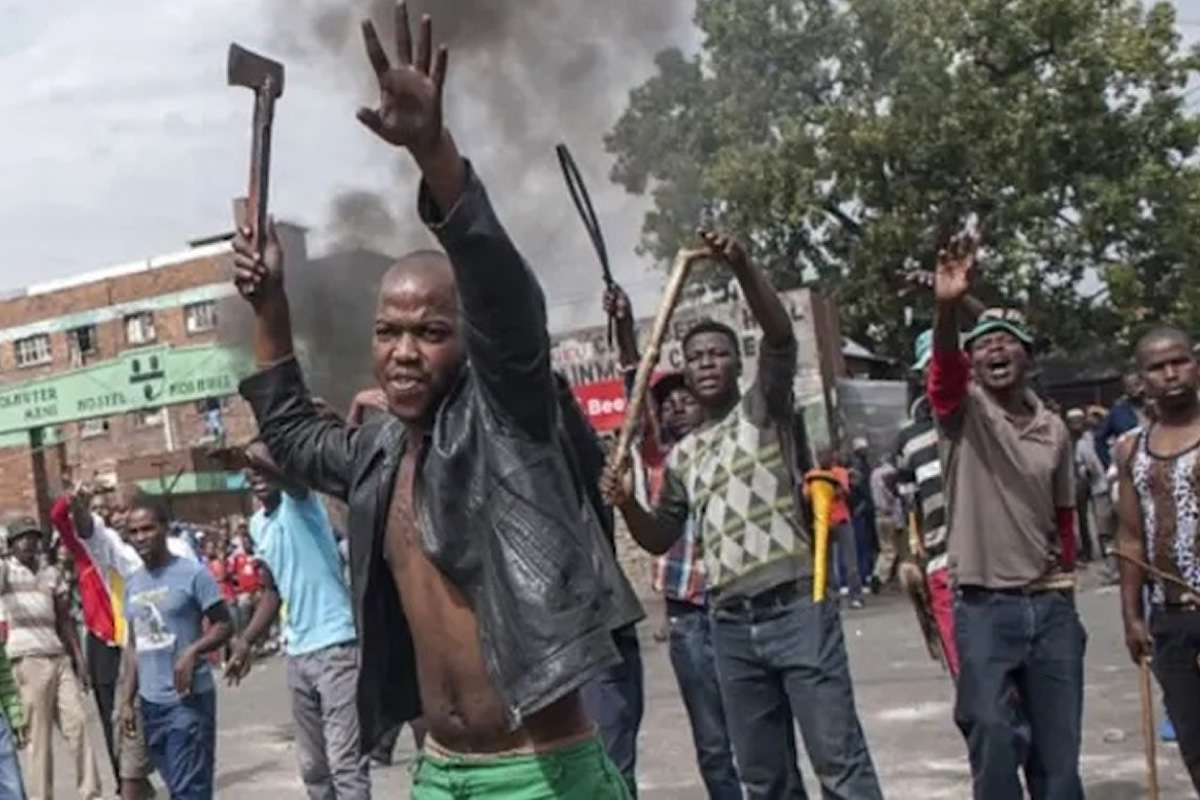 Group of men protesting in a street; a man in a black jacket raises a machete and his hand toward the camera while others hold sticks, with smoke rising in the background.