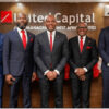 Five men in dark suits with red ties pose for a formal group photo in front of a United Capital sign in an office lobby.