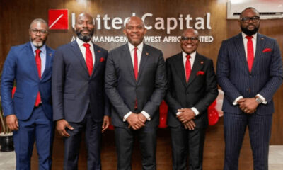 Five men in dark suits with red ties pose for a formal group photo in front of a United Capital sign in an office lobby.
