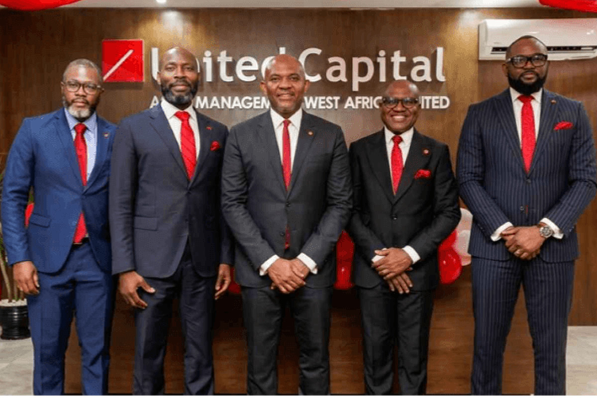 Five men in dark suits with red ties pose for a formal group photo in front of a United Capital sign in an office lobby.