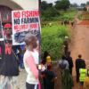 Protesters hold signs reading 'No Fishing, No Farming, No Livelihood' outside a building exterior.