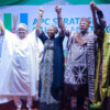 Group of diverse Nigerian politicians on stage, hands raised in unity at an APC rally with a branded backdrop.