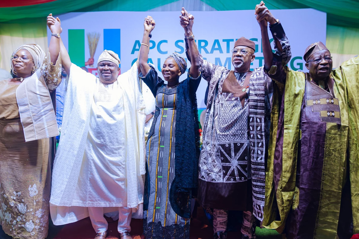 Group of diverse Nigerian politicians on stage, hands raised in unity at an APC rally with a branded backdrop.