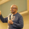 Man wearing glasses speaks into a handheld microphone while gesturing with his left hand in a conference setting.
