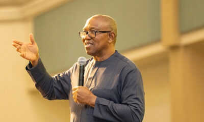 Man wearing glasses speaks into a handheld microphone while gesturing with his left hand in a conference setting.