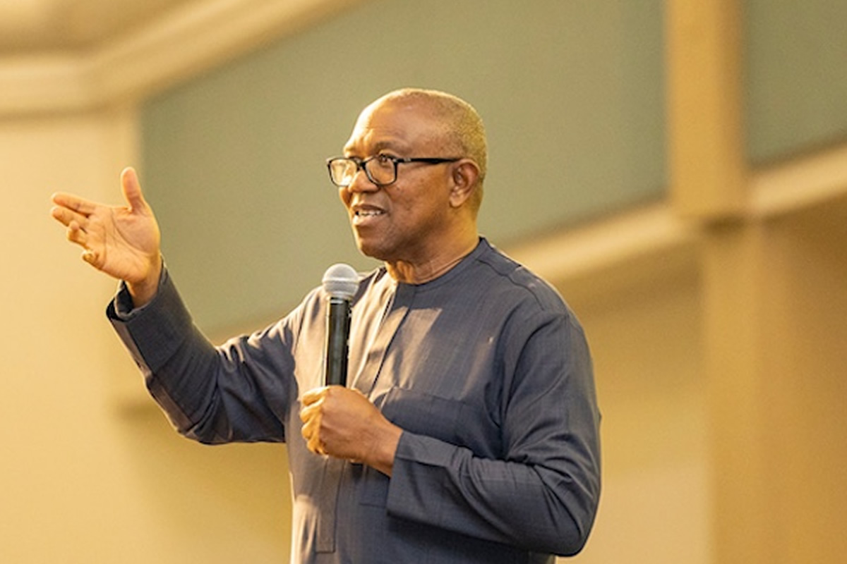 Man wearing glasses speaks into a handheld microphone while gesturing with his left hand in a conference setting.