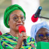 Woman in a bright green headwrap and patterned dress speaks into a red microphone at a public event.
