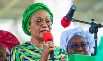 Woman in a bright green headwrap and patterned dress speaks into a red microphone at a public event.