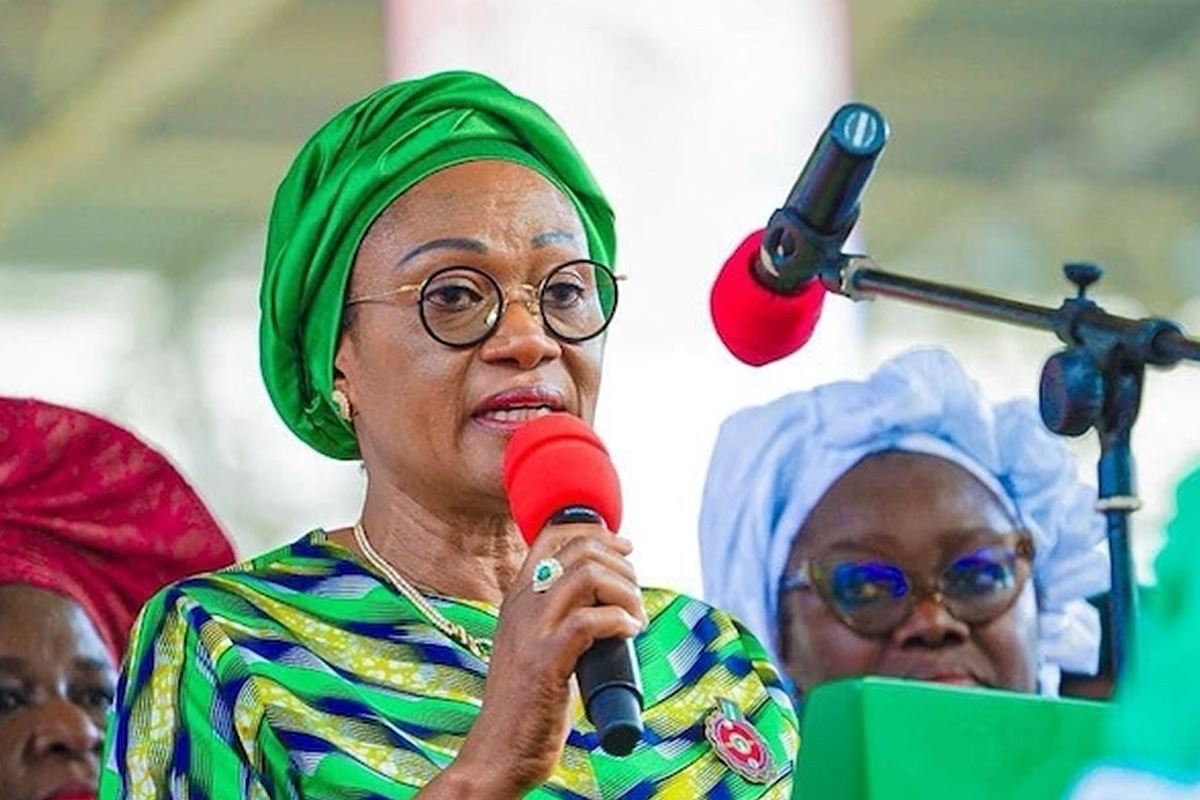 Woman in a bright green headwrap and patterned dress speaks into a red microphone at a public event.