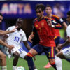 Soccer player in red jersey #7 dribbling the ball while four white-uniformed opponents close in on a green field.