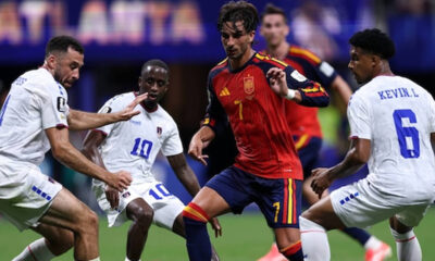 Soccer player in red jersey #7 dribbling the ball while four white-uniformed opponents close in on a green field.