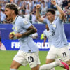 Two soccer players in light blue jerseys celebrate a goal on a packed stadium field, fists raised in triumph.