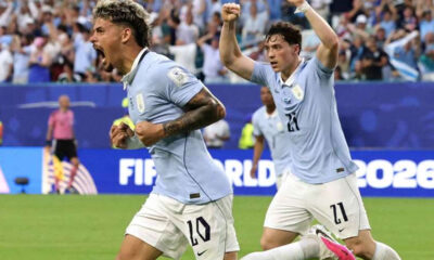 Two soccer players in light blue jerseys celebrate a goal on a packed stadium field, fists raised in triumph.