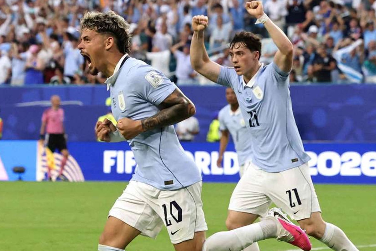 Two soccer players in light blue jerseys celebrate a goal on a packed stadium field, fists raised in triumph.