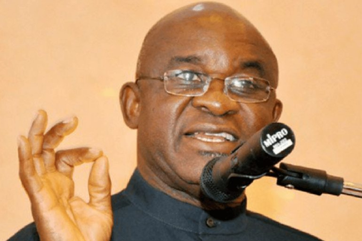 Man with glasses speaking into a microphone, making a small gesture with his right hand while addressing an audience.