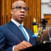 Man in a navy pinstripe suit, white shirt, and blue patterned tie speaks at a podium with a microphone in a formal setting, possibly a courtroom or conference room.