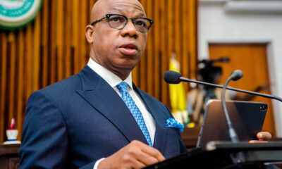 Man in a navy pinstripe suit, white shirt, and blue patterned tie speaks at a podium with a microphone in a formal setting, possibly a courtroom or conference room.