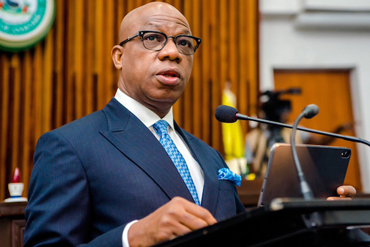 Man in a navy pinstripe suit, white shirt, and blue patterned tie speaks at a podium with a microphone in a formal setting, possibly a courtroom or conference room.