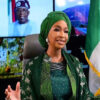 Nigerian woman in green traditional attire and gele, gesturing while seated in a studio with a Nigerian flag behind her.