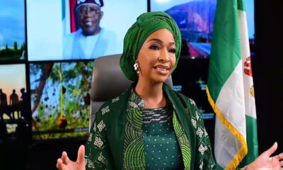 Nigerian woman in green traditional attire and gele, gesturing while seated in a studio with a Nigerian flag behind her.