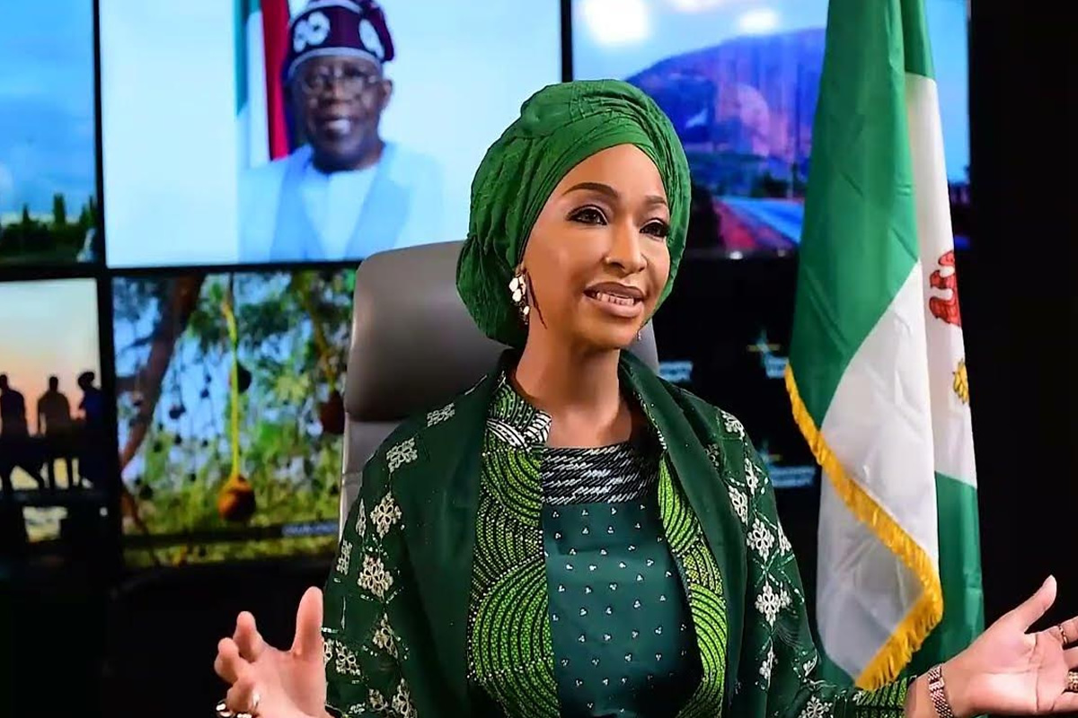 Nigerian woman in green traditional attire and gele, gesturing while seated in a studio with a Nigerian flag behind her.