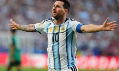 Argentine soccer player with arms outstretched celebrating on the field in a white and light-blue striped jersey with number 10, captain’s armband visible on the sleeve