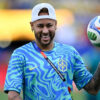 Brazilian footballer in a blue Brazil training jersey holding a colorful soccer ball, smiling on a field.