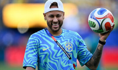 Brazilian footballer in a blue Brazil training jersey holding a colorful soccer ball, smiling on a field.